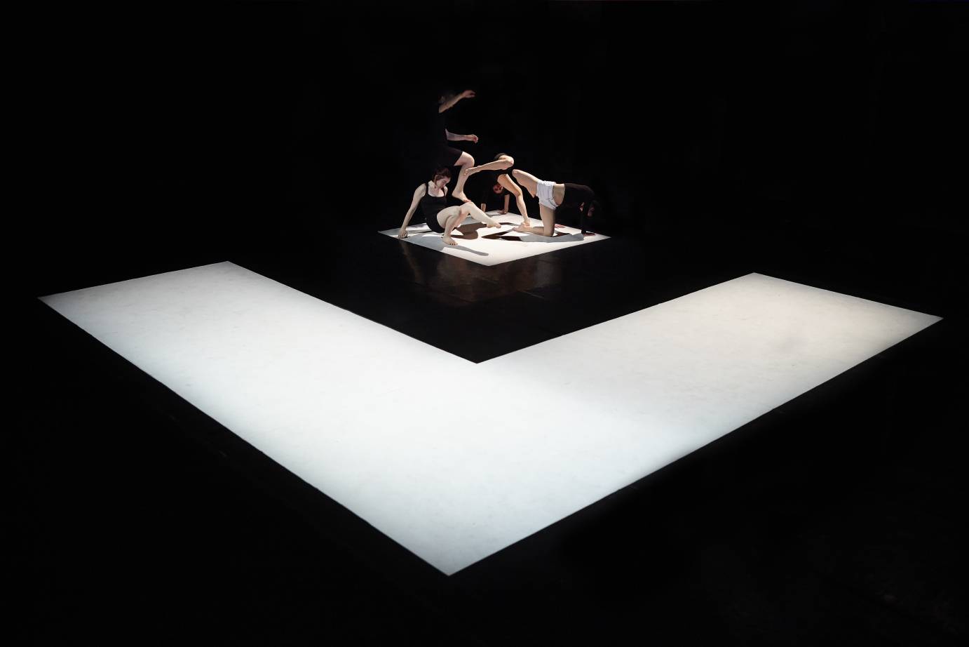 On a floor of a big white square holding a small black one, performers crouch with a leg or arm extended 